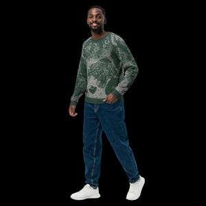 K&ouml;nnte beinhalten: Ein gr&uuml;n-grau gemustertes Sweatshirt. Das Sweatshirt hat ein einzigartiges Design mit einer Mischung aus Gr&uuml;n- und Graut&ouml;nen. Die Person tr&auml;gt eine blaue Jeans und wei&szlig;e Turnschuhe. Der Hintergrund ist schwarz.