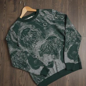 K&ouml;nnte beinhalten: Gr&uuml;ner und grauer Rundhals-Pullover mit Quallenmuster. Der Pullover hat einen dunkelgr&uuml;nen gerippten Kragen, B&uuml;ndchen und Saum. Das Quallen-Design ist in einer helleren grauen Farbe gehalten und bildet einen Kontrast zum gr&uuml;nen Hintergrund.