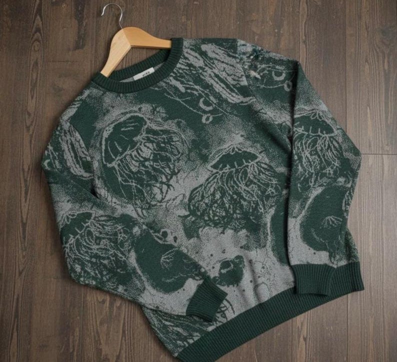 K&ouml;nnte beinhalten: Gr&uuml;ner und grauer Rundhals-Pullover mit Quallenmuster. Der Pullover hat einen dunkelgr&uuml;nen gerippten Kragen, B&uuml;ndchen und Saum. Das Quallen-Design ist in einer helleren grauen Farbe gehalten und bildet einen Kontrast zum gr&uuml;nen Hintergrund.