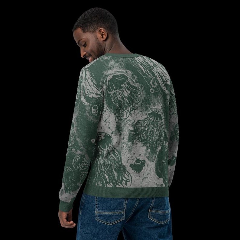 K&ouml;nnte beinhalten: Ein gr&uuml;ner Rundhals-Pullover mit einem sich wiederholenden Muster aus Quallen in Grau- und Wei&szlig;t&ouml;nen. Der Pullover ist aus einem weichen Material gefertigt und wird von einer Person getragen, die blaue Jeans tr&auml;gt.