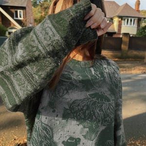 K&ouml;nnte beinhalten: Ein gr&uuml;n-grau gemusterter Pullover. Der Pullover hat einen Rundhalsausschnitt und lange &Auml;rmel. Das Muster scheint ein florales Design zu sein. Der Pullover ist aus einem weichen, gestrickten Material gefertigt. Die &Auml;rmel sind hochgezogen, um die Hand zu bedecken.
