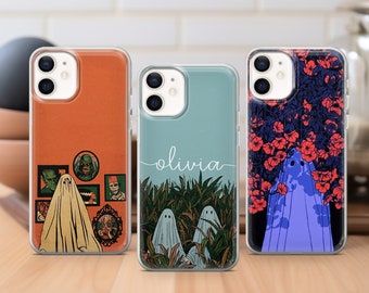 May include: Three clear phone cases with different designs. The first case has a red, orange, and white design with a ghost and other spooky characters. The second case has a blue and green design with a ghost and the word "olivia" written in white. The third case has a black and white design with a ghost and red flowers.