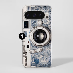Pu&ograve; includere: Una custodia per telefono con un design di fotocamera vintage. La custodia presenta un motivo floreale bianco e blu con un obiettivo della fotocamera dettagliato ed elementi meccanici. Il design ricorda le classiche attrezzature fotografiche.