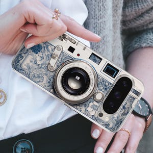 Pu&ograve; includere: Una custodia per telefono con un design a fotocamera. La custodia presenta un motivo floreale bianco e blu, con un obiettivo e pulsanti. La custodia &egrave; tenuta da una persona che indossa un anello.