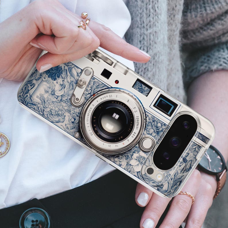 Pu&ograve; includere: Una custodia per telefono con un design a fotocamera. La custodia presenta un motivo floreale bianco e blu, con un obiettivo e pulsanti. La custodia &egrave; tenuta da una persona che indossa un anello.