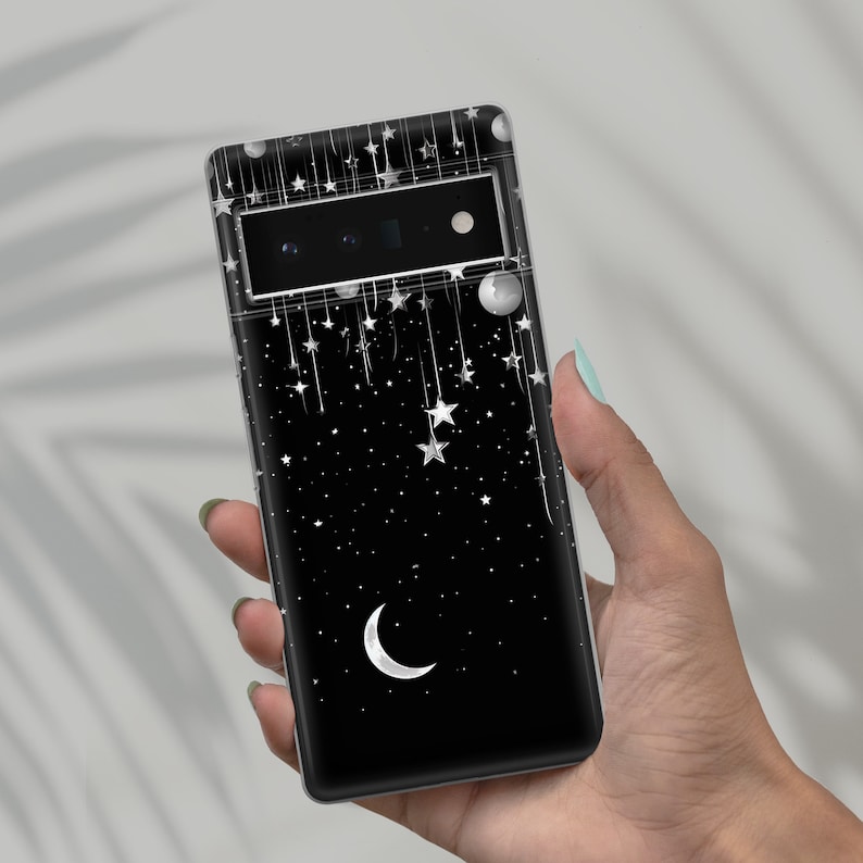 Puede incluir: Una funda de tel&eacute;fono negra con un dise&ntilde;o de noche estrellada. La funda presenta una luna creciente, estrellas y luces de cadena blancas.