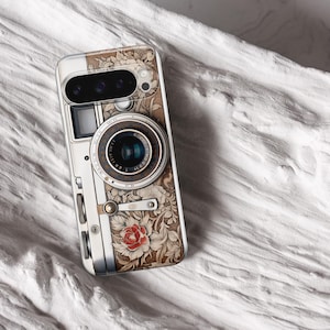 Pu&ograve; includere: Una custodia per telefono con un design di fotocamera vintage. La custodia presenta un motivo floreale dettagliato nei toni del marrone e del crema, con un accento di rosa rossa. L'obiettivo e i pulsanti della fotocamera sono rappresentati in modo realistico.