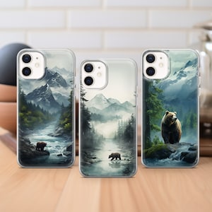 May include: Three clear phone cases with a mountain and river landscape design. Each case features a brown bear in a different pose. The cases are for iPhone 13 Pro.