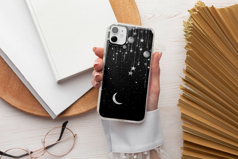 May include: A clear phone case with a black background and a white crescent moon, stars, and dangling ornaments.