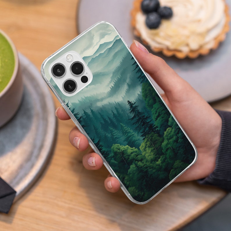 May include: Clear phone case featuring a landscape design of a forest with green trees and mountains. The case has a clear border and covers the back of the phone. The design has a watercolor effect.