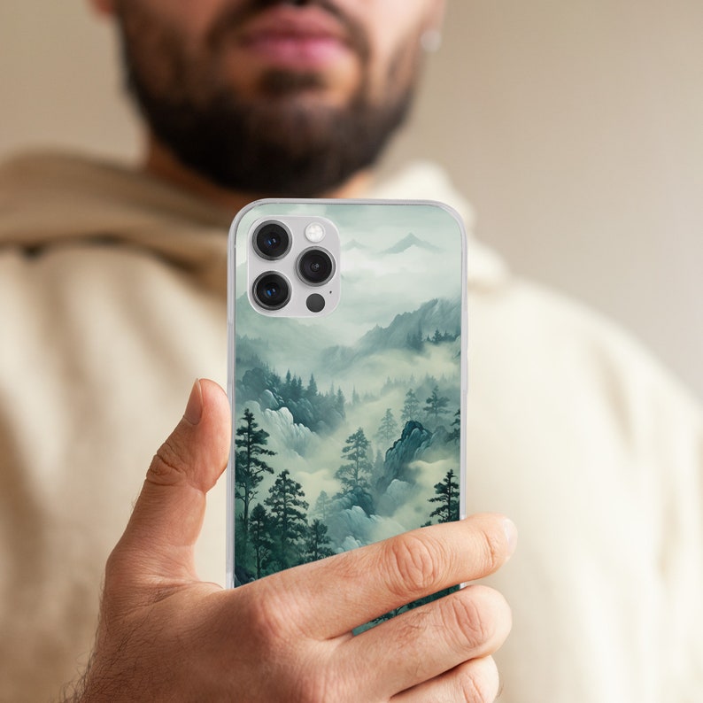 May include: A clear phone case featuring a misty mountain landscape design. The artwork depicts a serene scene with evergreen trees, rolling hills, and a soft, atmospheric fog in shades of green and gray.