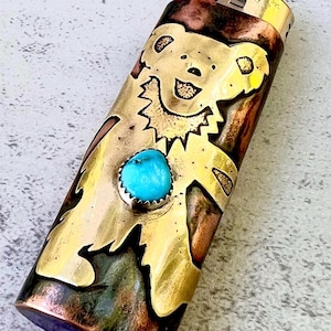 May include: A decorative lighter with a brass bear design and a turquoise stone. The lighter has a red button and a metallic finish. The bear is a stylized design with a smiling face.