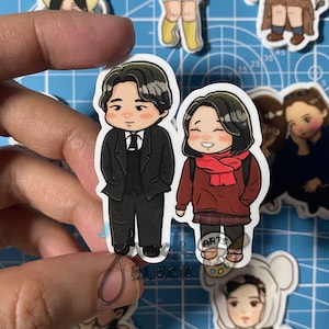 May include: Two cartoon stickers of a man and woman. The man is wearing a black suit and tie. The woman is wearing a red sweater, a red scarf, and a plaid skirt. The stickers are on a blue and white grid background.