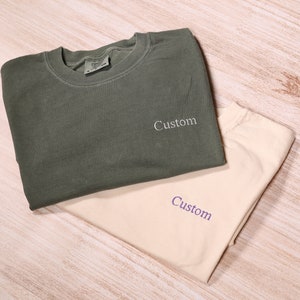 May include: Two t-shirts, one olive green and one beige, both folded and stacked on top of each other. The shirts have the word "Custom" embroidered on them.