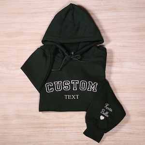 May include: Dark green hooded sweatshirt with white text reading "CUSTOM" and "TEXT". The sleeve features the names "Kevin Bella" and a heart symbol. The sweatshirt is folded on a light wood grain surface.