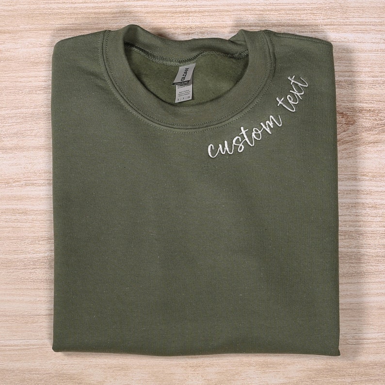 May include: A folded olive green sweatshirt with the text "custom text" embroidered on the front.