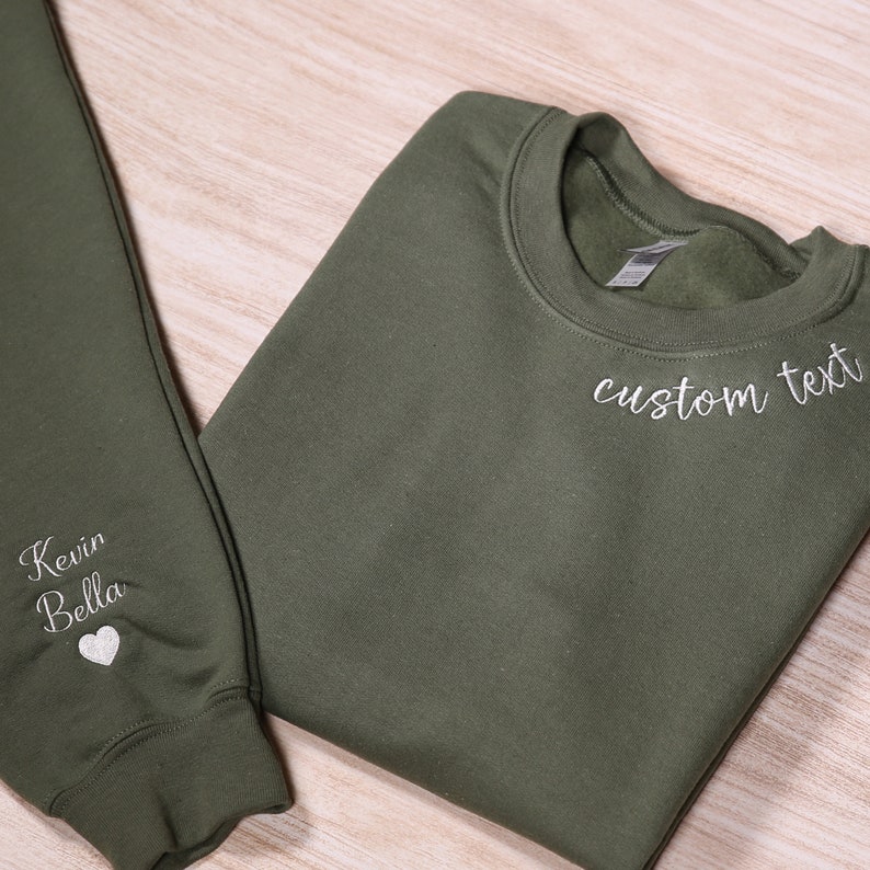 May include: A green sweatshirt and sweatpants set with white embroidery. The sweatshirt has "custom text" embroidered on the front and the sweatpants have "Kevin Bella" and a heart embroidered on the leg.