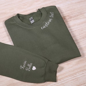 May include: A green sweatshirt with white embroidery. The text "custom text" is embroidered on the neckline. The text "Kevin Bella" and a heart are embroidered on the sleeve.