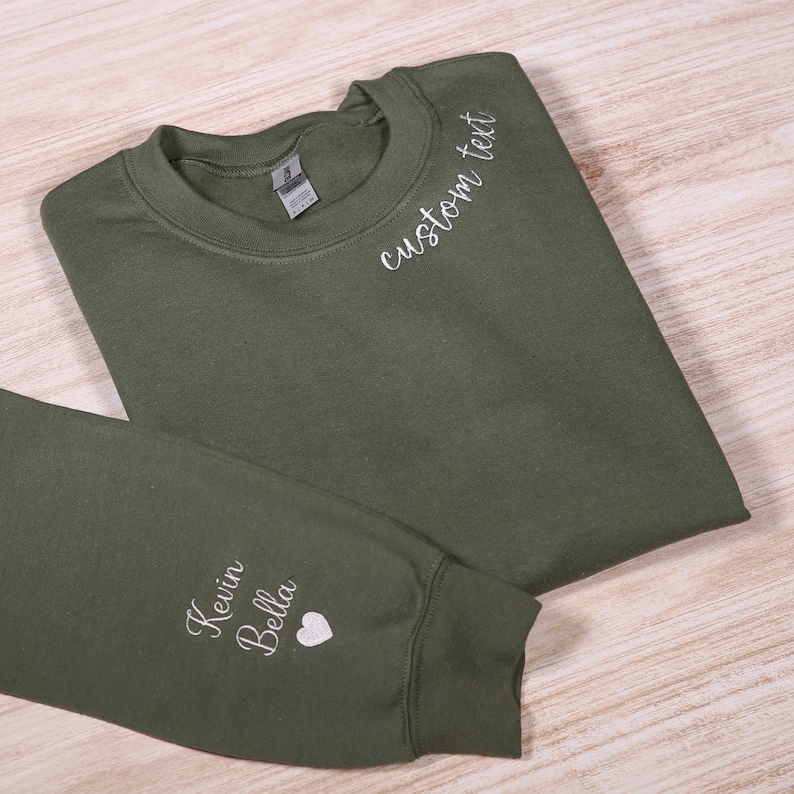 May include: A green sweatshirt with white embroidery. The text "custom text" is embroidered on the neckline. The text "Kevin Bella" and a heart are embroidered on the sleeve.