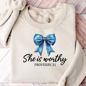 May include: A beige sweatshirt with a blue denim bow and the text "She is worthy Proverbs 31" printed on the front.
