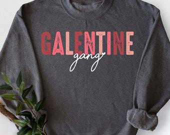 May include: Dark gray sweatshirt with the text "GALENTINE gang" in red and pink letters. The word "gang" is in a handwritten font. A sprig of greenery is draped on the bottom left.