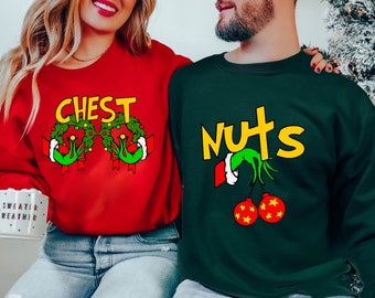 Puede incluir: Sudaderas navideñas rojas y verdes. Una sudadera roja con la palabra "CHEST" en amarillo, decorada con coronas verdes. La otra es verde oscuro con la palabra "NUTS" en amarillo, con manos tipo Grinch sosteniendo adornos rojos.