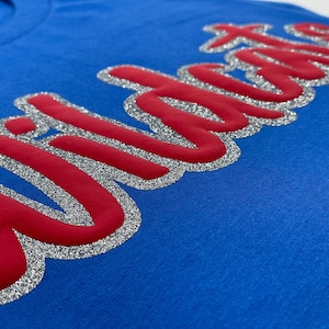May include: Close-up of a blue t-shirt with the word "Wildcats" in red, outlined with silver glitter. The text is in a cursive font and is slightly raised from the fabric.