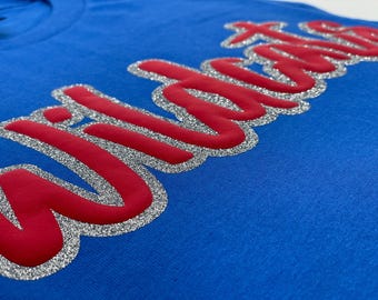 Pode incluir: Close-up de uma camiseta azul com a palavra "Wildcats" em vermelho, contornada com glitter prateado. O texto está em uma fonte cursiva e ligeiramente em relevo no tecido.