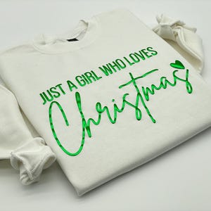 May include: Cream-colored sweatshirt with the text "JUST A GIRL WHO LOVES Christmas" in bright green script. The long sleeves are folded, and a small green heart is next to the word "Christmas". A festive holiday-themed apparel item.