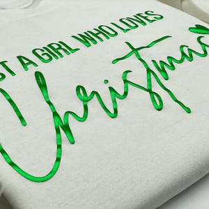 May include: Off-white sweatshirt with the text "JUST A GIRL WHO LOVES Christmas" in bright green cursive lettering. The sweatshirt has a soft, textured appearance and a casual style, perfect for the holiday season.