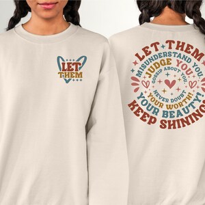 May include: A beige sweatshirt with a graphic on the front that says "Let Them" in a heart shape and a graphic on the back that says "Let Them Misunderstand You. Judge You. Gossip About You. Never Doubt Your Worth. Your Beauty! Keep Shining!" in a circle with stars and hearts.