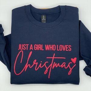 May include: A navy blue sweatshirt with the text "JUST A GIRL WHO LOVES Christmas" in red script. A red heart is next to the word "Christmas". The sweatshirt is folded neatly.