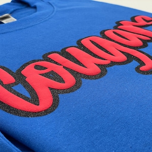 May include: A royal blue sweatshirt with the word "Cougars" in large, red, cursive lettering outlined in black glitter. The sweatshirt has a crew neck and is made of a soft, textured fabric.