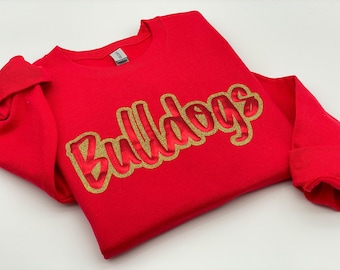 Pode incluir: Um moletom vermelho com a palavra "Bulldogs" em letras douradas com glitter. O moletom é feito de um material macio e tem mangas compridas. A palavra "Bulldogs" está em uma fonte ousada e estilizada.