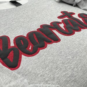 May include: A heather gray sweatshirt with the word "Beatles" in large, stylized lettering across the front. The text is black with a red glitter outline, creating a bold contrast against the gray fabric. The sweatshirt has a crew neck and long sleeves.