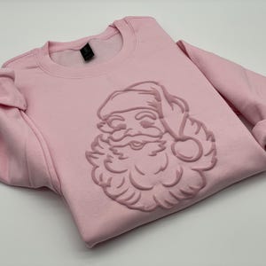 May include: A light pink sweatshirt featuring a raised Santa Claus design on the front. The long-sleeved top has a crew neck and folded cuffs. The Santa Claus design is in a matching pink color.