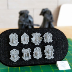 May include: Eight grey metal decorative plaques with different designs, including a skull, a chalice, and a winged figure, displayed on a black circular tray.