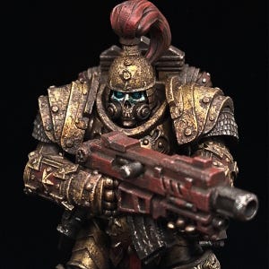 May include: A miniature figure of a space marine wearing gold and red armour, holding a red and black gun. The figure has a skull face mask and blue eyes.