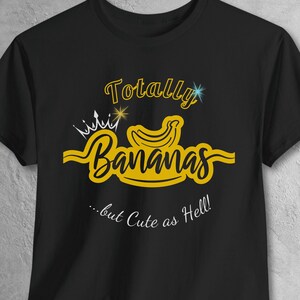May include: Black t-shirt with a yellow graphic that reads "Totally Bananas" with a banana and crown design. The text "...but Cute as Hell!" is below the main text. The shirt is made of a soft material.