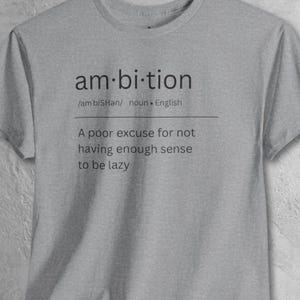 May include: A heather gray t-shirt with the word "ambition" printed in black, along with its pronunciation and definition. Below the word is the phrase "A poor excuse for not having enough sense to be lazy."