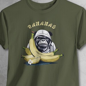 May include: Olive green t-shirt with a graphic of a gorilla wearing a white hat and shirt, surrounded by bananas. The word "BANANAS" is arched above the gorilla. The bananas are yellow with green stems and a small white flower.