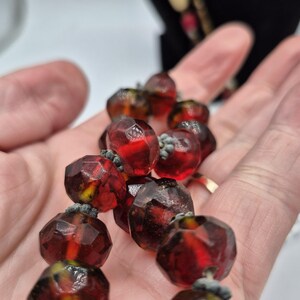 May include: A necklace featuring faceted, translucent red and amber-coloured beads. The beads are round with multiple flat surfaces and are strung together with small, dark grey, textured spacers between each individual bead in the strand.