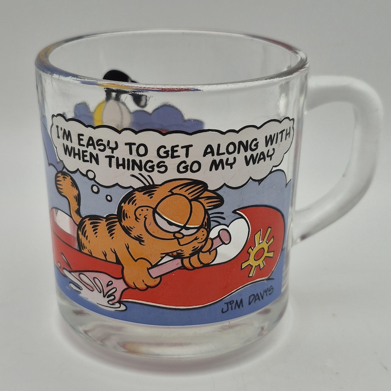 May include: Clear glass mug featuring Garfield the cat on a red raft with the text "I'M EASY TO GET ALONG WITH WHEN THINGS GO MY WAY." The mug has a white handle and a cartoon illustration.