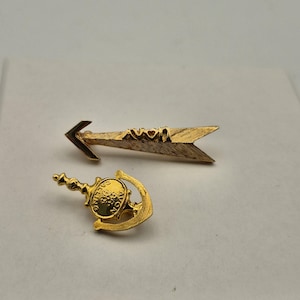 May include: Two gold-coloured Avon pins. One pin is an arrow with the word "AVON" etched into it. The other pin is a gold-coloured anchor with the word "AVON" and a floral design.