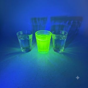 May include: Three clear glass shot glasses arranged in a row. The centre glass glows with a bright, vibrant neon green light, while the two glasses on either side remain clear and transparent under the blue ambient lighting conditions.