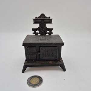 May include: A small, dark brown cast iron stove with the word "DOT" on the front. It has a decorative top and legs. A coin is in the foreground. The stove is likely a miniature model.