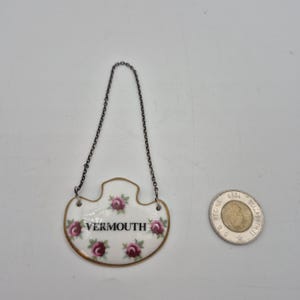 May include: A white porcelain vermouth label with a gold trim and a silver chain. The label is decorated with pink roses and the word "VERMOUTH" in black lettering.