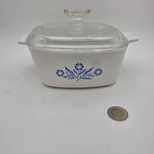 May include: A white rectangular casserole dish with integrated side handles and a clear glass lid. The front of the dish features a blue floral pattern consisting of three stylised flowers and leaves. A small circular coin is placed nearby for scale.