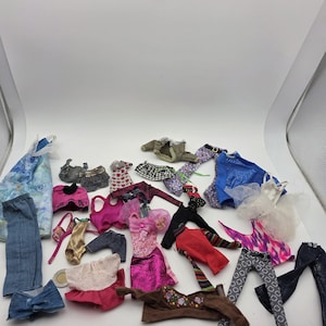 Op de afbeelding: Een verzameling diverse poppenkleding, waaronder verschillende tops, broeken, rokken en jurken in meerdere kleuren, patronen en texturen zoals denim, metallic stof, bloemenprints en tule, verspreid over een effen wit oppervlak.