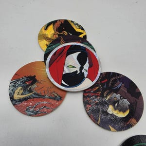 May include: A collection of five circular pogs featuring colorful comic book illustrations. The designs include a masked character with a red hood, a creature with sharp teeth, and various abstract patterns in shades of red, yellow, and black.